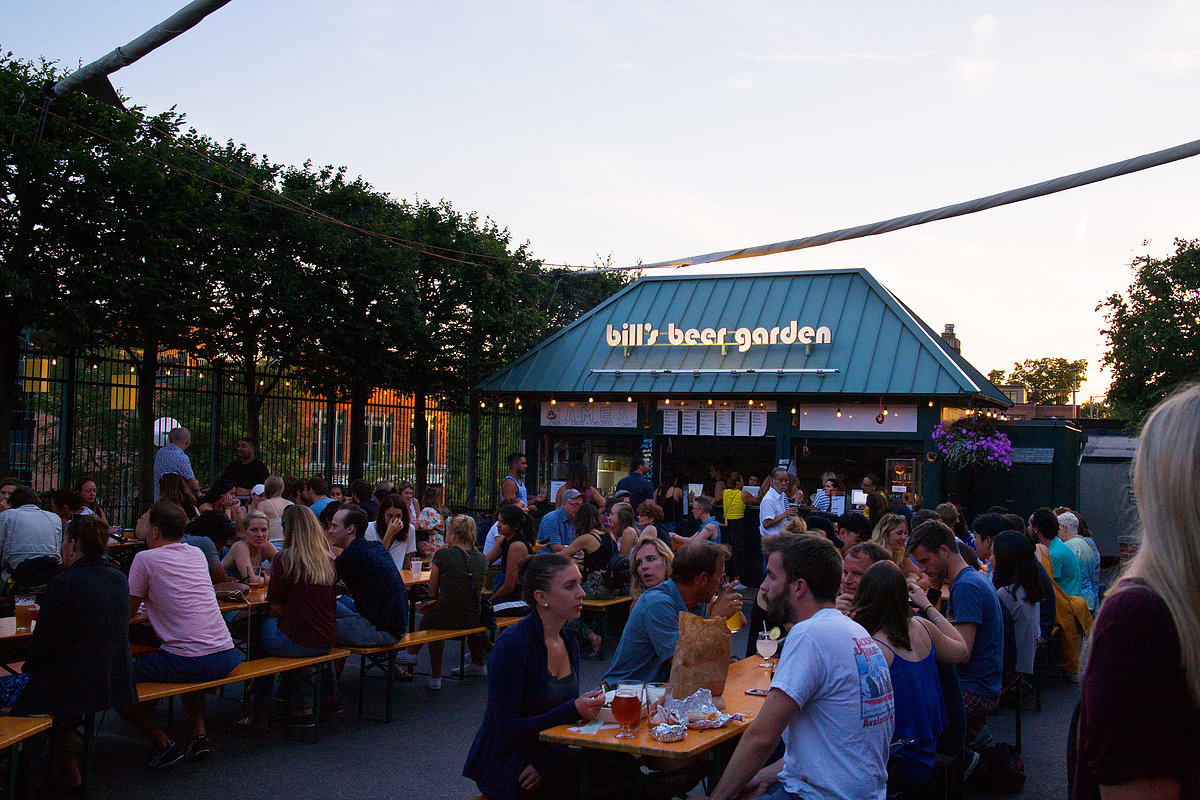people sitting outdoors at wooden communal tables at twilight. There’s a Bill Beer Garden sign on a blue roof in the background.