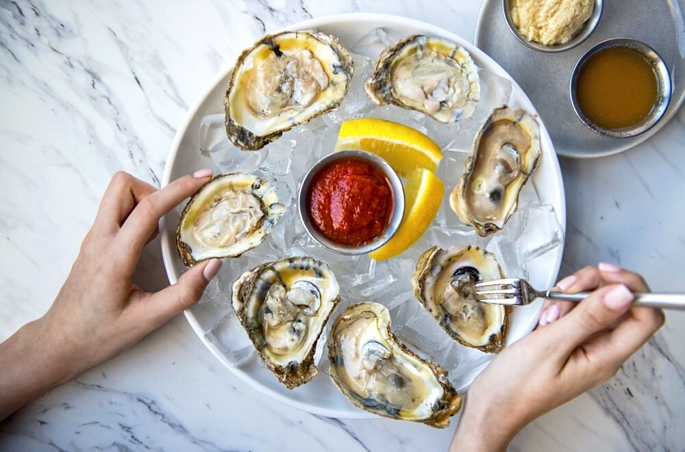 A bunch of oysters on a platter with red sauce and lemon slices at the center. A hand picks up one and holds a shrimp fork in the other.