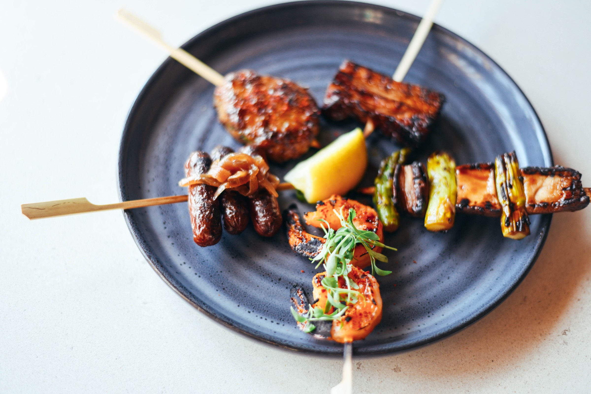 Skewers of meat and vegetables from Ima Izakaya in Corktown, Detroit, Michigan set on a blue round plate on a white surface.