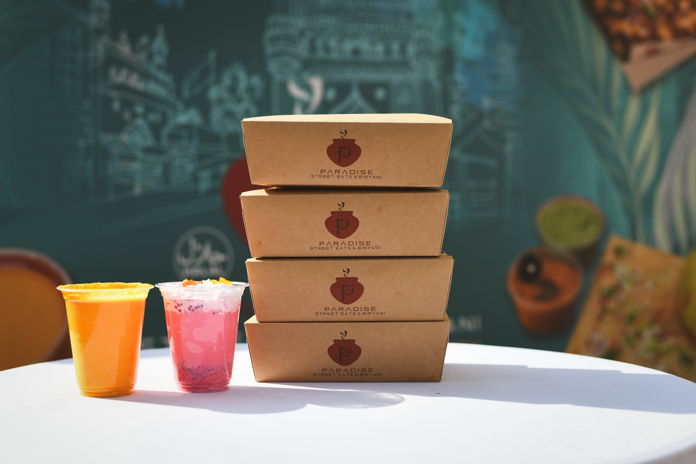 Four brown cardboard boxes that say Paradise Street Eats and two cups, one with orange and one with pink substance inside them, set on a white surface in front of a turquoise colored food truck in Detroit, Michigan.