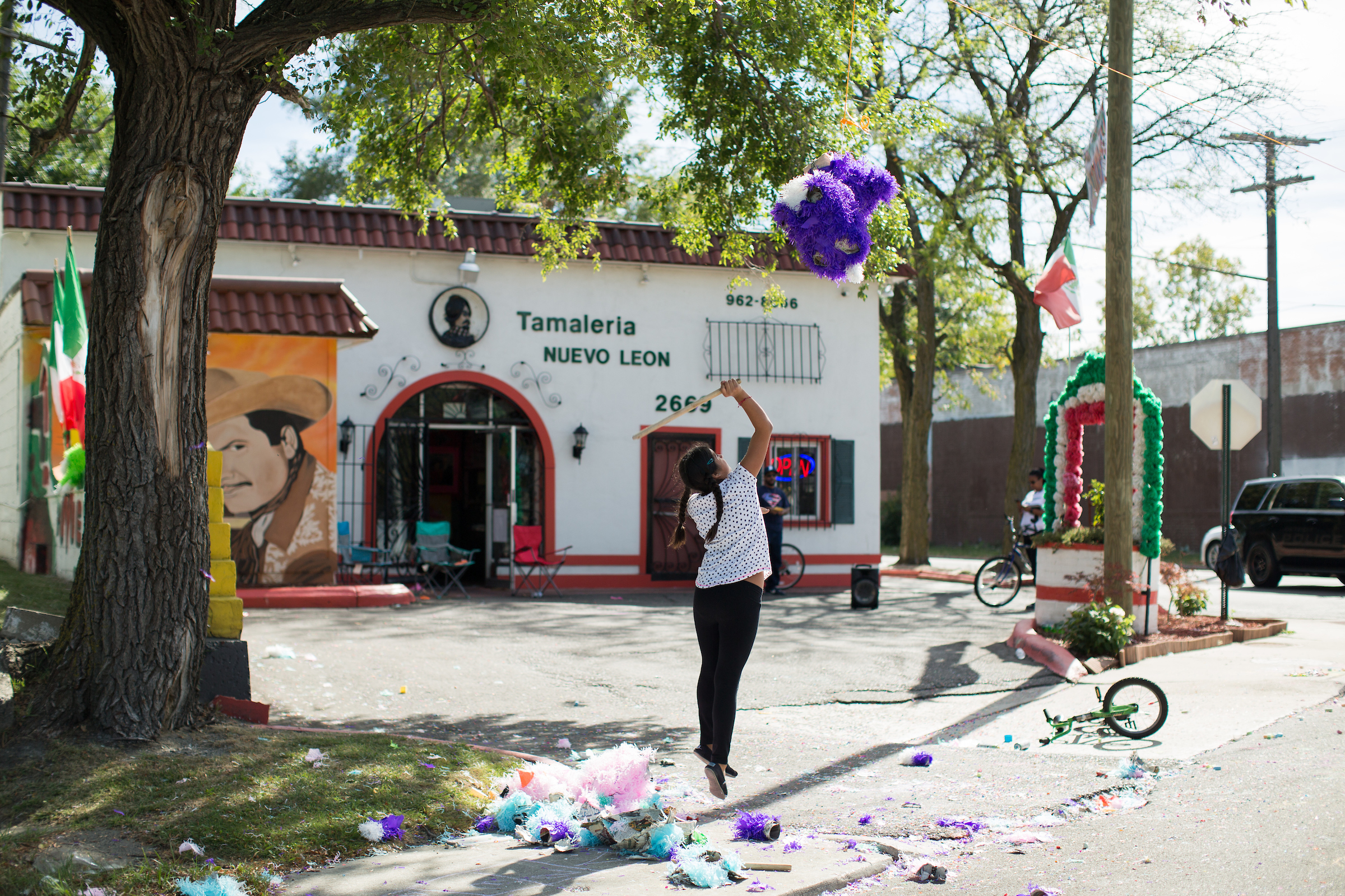 A girl in a white shirt and black jeans is shown mid-air jumping to hit a purple piñata in front of the white and red Tamaleria Nuevo Leon restaurant.