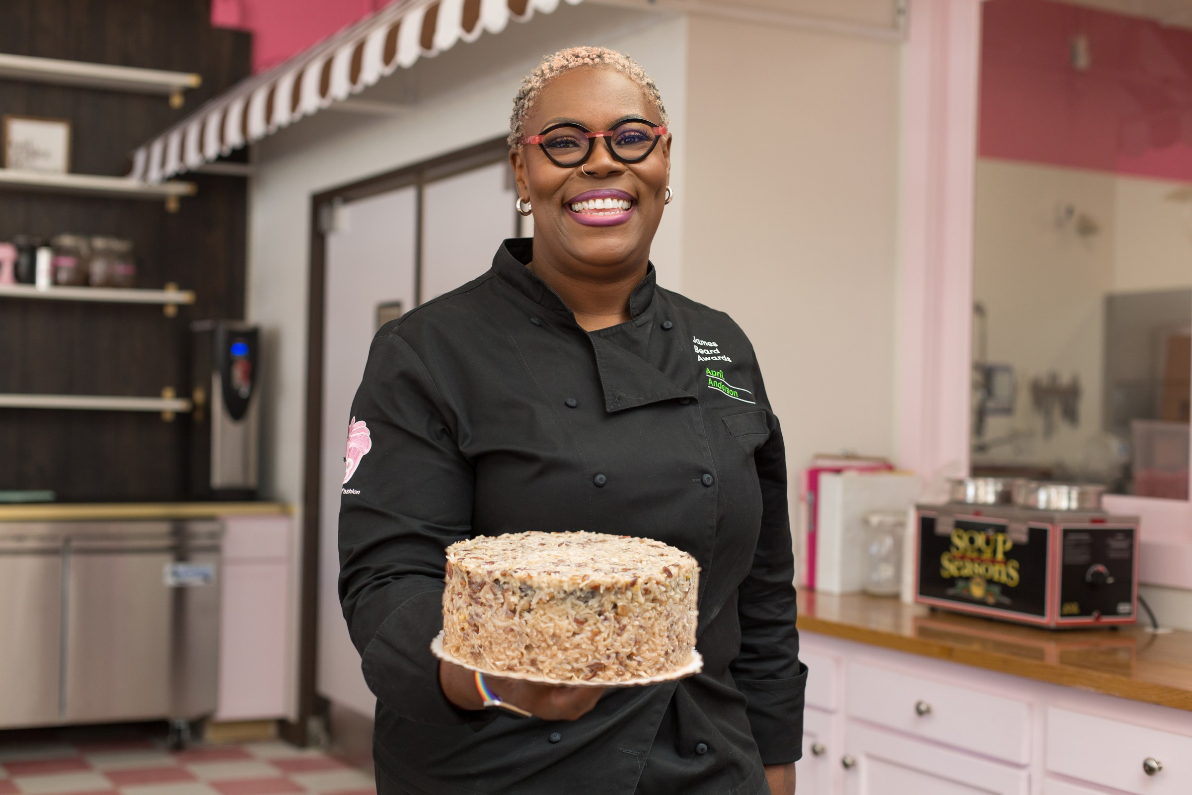 A woman with glasses, a black chef’s jacket, smiling, holding a round cake in a shop decorated in pinks, blacks, and whites.