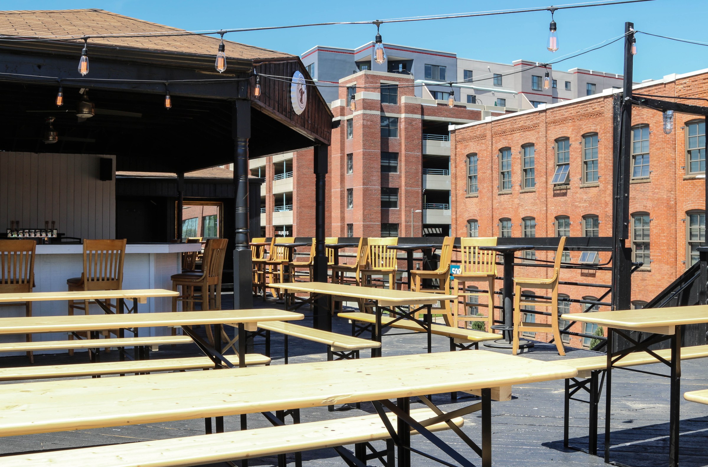 Several long wooden picnic tables accompanied by benches atop a rooftop that has a bar with chairs in the background. Two midrise buildings are in view.