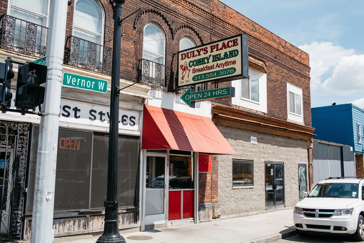 The exterior of Duly’s Place is shown in an old brick building next to barbershop Southwest Styles. Duly’s has a green and red sign with an arrow pointing towards the restaurant that says “24 Hours” and a red awning.