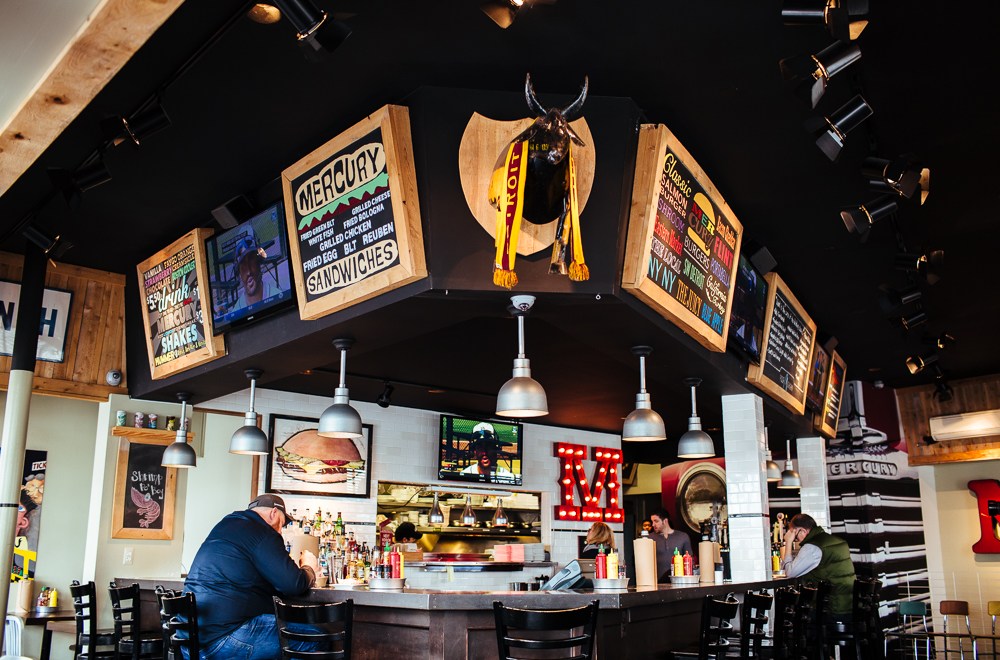 The interior of Mercury Burger & Bar with signs above a bar, and a person sitting at the bar to the left.