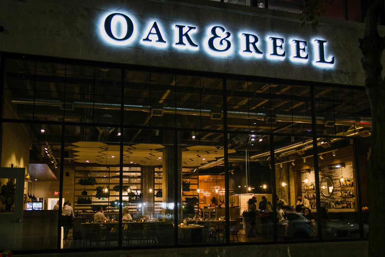 Diners are viewed through large exterior windows beneath the sign for Oak and Reel at night.